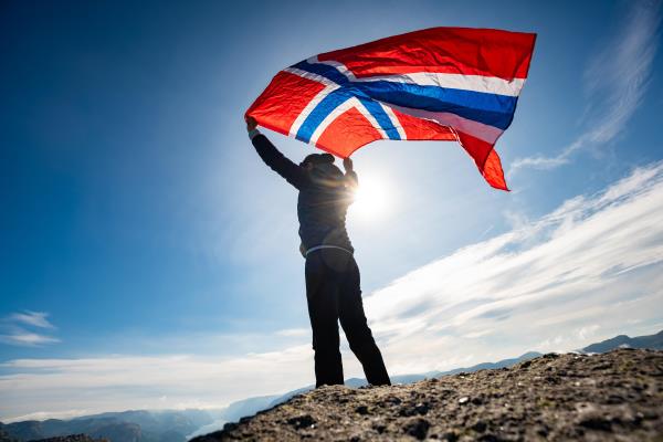 En person som står på toppen av et fjell og holder et stort norgesflagg over hodet - Klikk for stort bilde