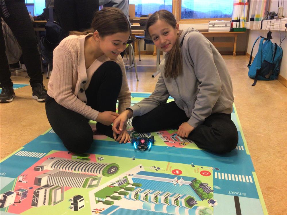 To jenter sitter på gulvet i klasserommet og arbeider med koding av robot - Klikk for stort bilde