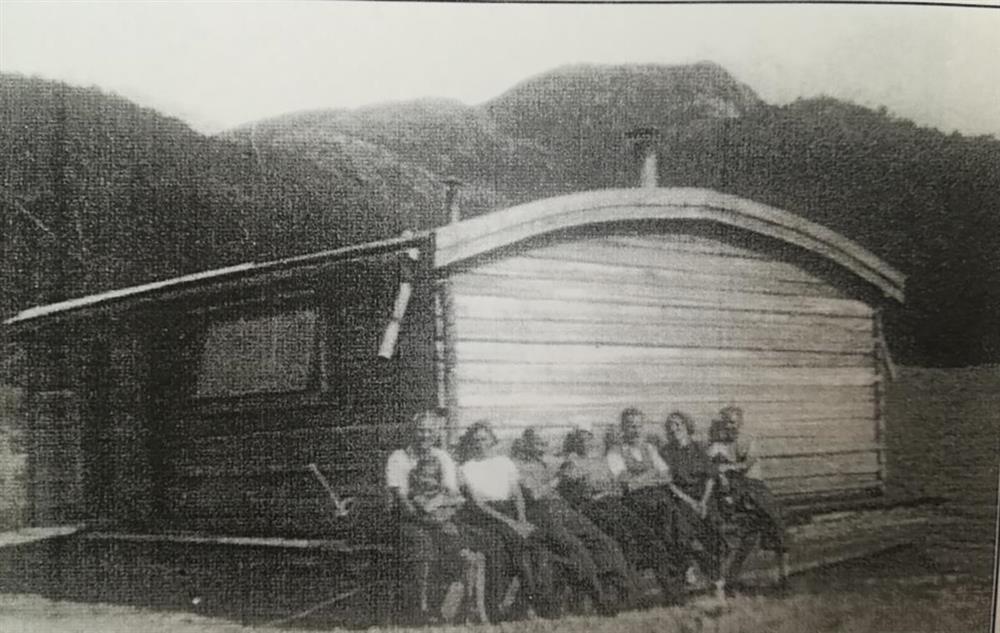 Skogbruksvirksomhet. Gammelt, avfotografert bilde av en liten hytte med mennesker som sitter på benken foran. - Klikk for stort bilde
